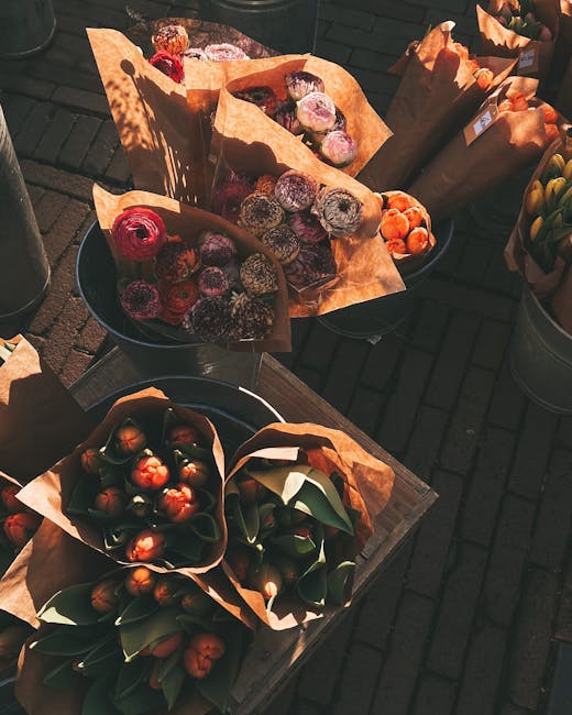 A collection of floral bouquets and flower arrangements displayed outdoors on dark wooden tables. The bouquets are wrapped in brown kraft paper and arranged in round black buckets, featuring an array