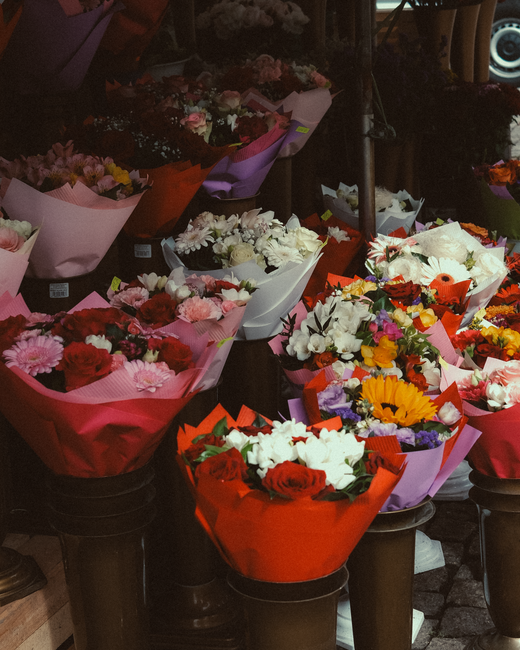 An array of fresh floral bouquets displayed outdoors under low light, arranged in colorful wrapping paper including red, purple, and white, with a variety of flowers such as roses, daisies, lilies, an