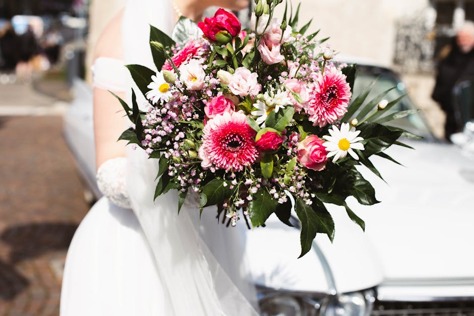 A close-up of a bridal bouquet featuring vibrant pink gerbera daisies, soft pink roses, white daisies with yellow centers, and delicate pink baby's breath, all arranged with lush green foliage. The bo