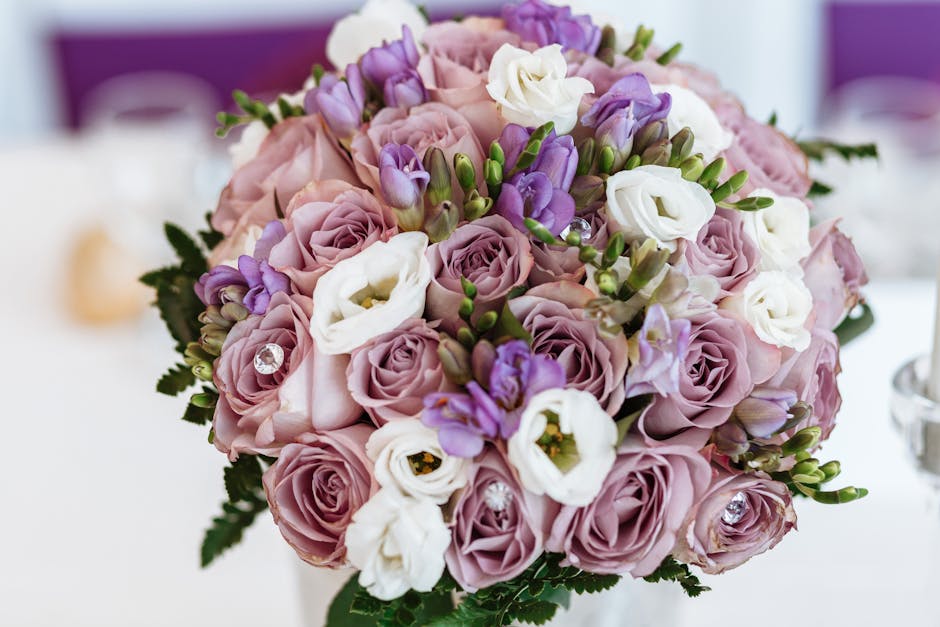 A close-up view of a floral bouquet featuring soft pink roses, white lisianthus, and purple freesias, arranged in a dense, rounded style. The flowers showcase fresh, delicate petals with a velvety tex