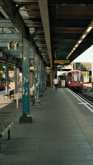 An outdoor view of Willesden Green station platform with a blue-painted metal canopy structure, showing signs of weathered paint and rust. A red train is arriving or departing on the tracks, while a s