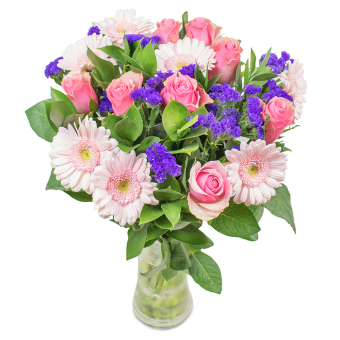 Arranged with care against a pristine backdrop, this vibrant floral bouquet in a clear glass vase is a joyful nod to Willesden's spirited neighbourhoods. The centerpiece displays about seven to nine medium-pink roses, some delicately furled and others just beginning to unfurl, each with perfectly spiraled petals hinting at timeless affection. Five to seven pale pink gerbera daisies, marked by their broad, sun-like faces and almost silvery centers, echo the fresh light that fills the area's open parks. Clusters of vivid purple statice are interspersed among the main stems, providing dynamic contrast and a lively punch of colour evocative of Willesden's multicultural vibrancy. Beneath, a lush foundation of deep green foliage spills gracefully, reminiscent of morning walks down Harlesden Road after spring rain. The transparent vase hints at freshly delivered beauty from Flower Delivery Willesden, with water and stems visible for an authentic touch. Suitable for occasions from birthdays to anniversaries or simply to brighten a workspace in Willesden Green, this bouquet embodies feelings of joy, celebration, and a sense of heartfelt belonging in North West London.